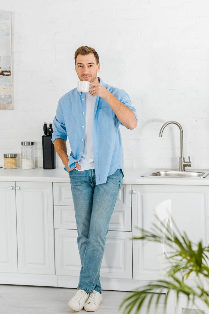 jóképű férfi alkalmi ruhák, látszó-on fényképezőgép, és kávét iszik a konyhában - Fotó, kép