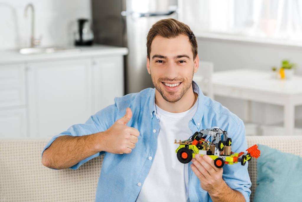 handsome smiling man looking at camera, holding toy car and showing thumb up sign at home - Photo, Image