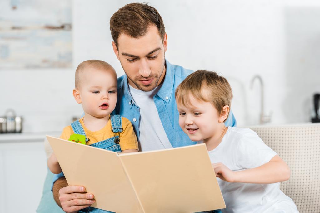 przystojny ojciec siedzi i czytanie książki przedszkolak i maluch synom w domu - Zdjęcie, obraz