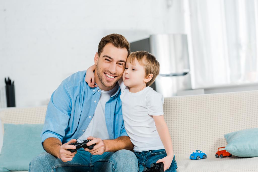 Дошкольный сын с джойстиком обнимает улыбающегося отца во время видеоигры дома
  - Фото, изображение