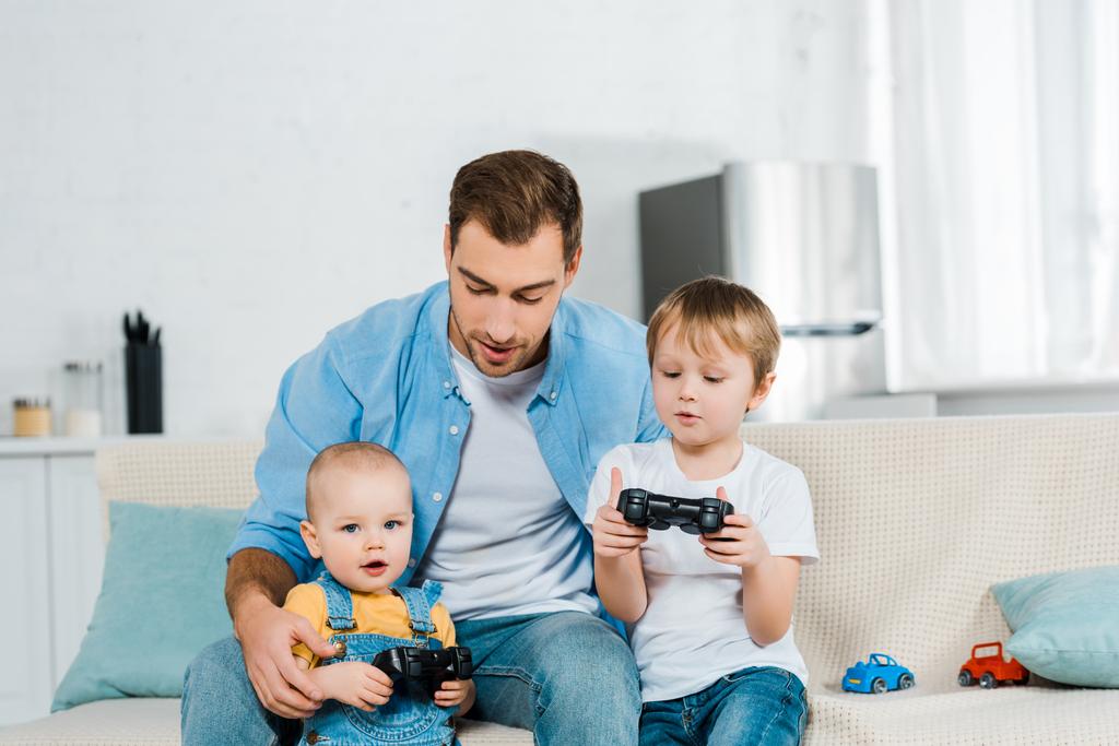 father playing video game with preschooler and toddler sons while sitting on couch at home - Photo, Image