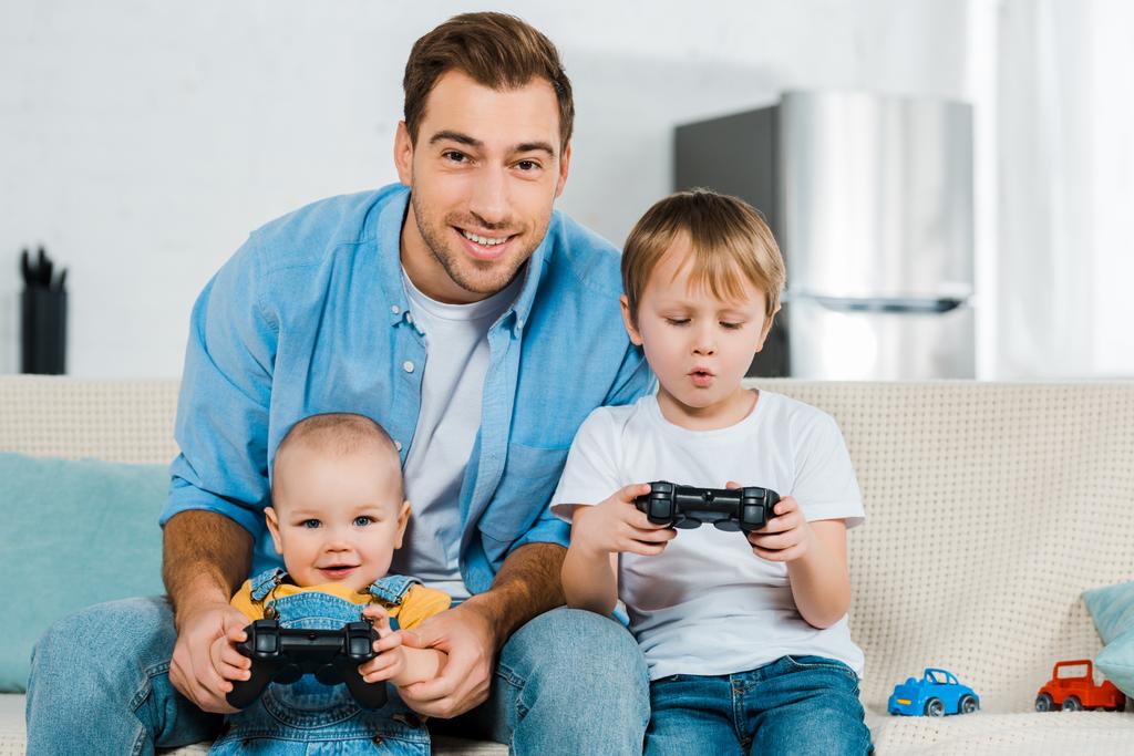 padre sonriente jugando videojuego con niños preescolares y niños pequeños adorables mientras está sentado en el sofá en casa
 - Foto, imagen