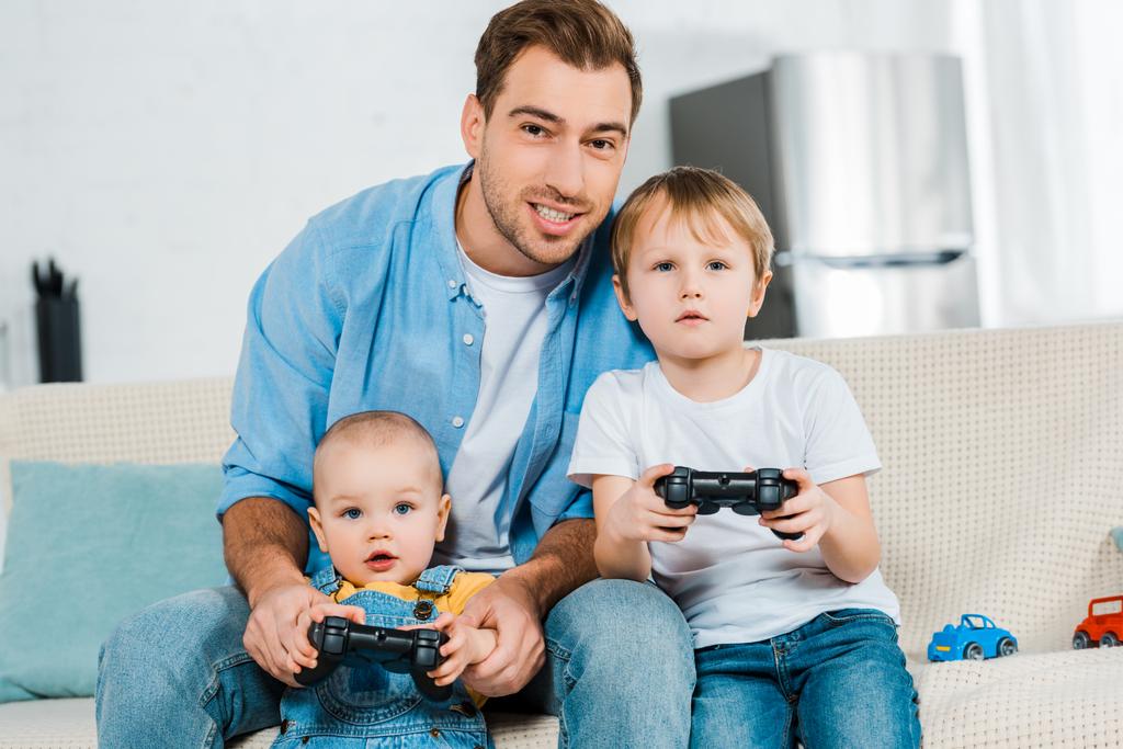 smiling father playing video game with preschooler and toddler sons while sitting on couch at home - Photo, Image