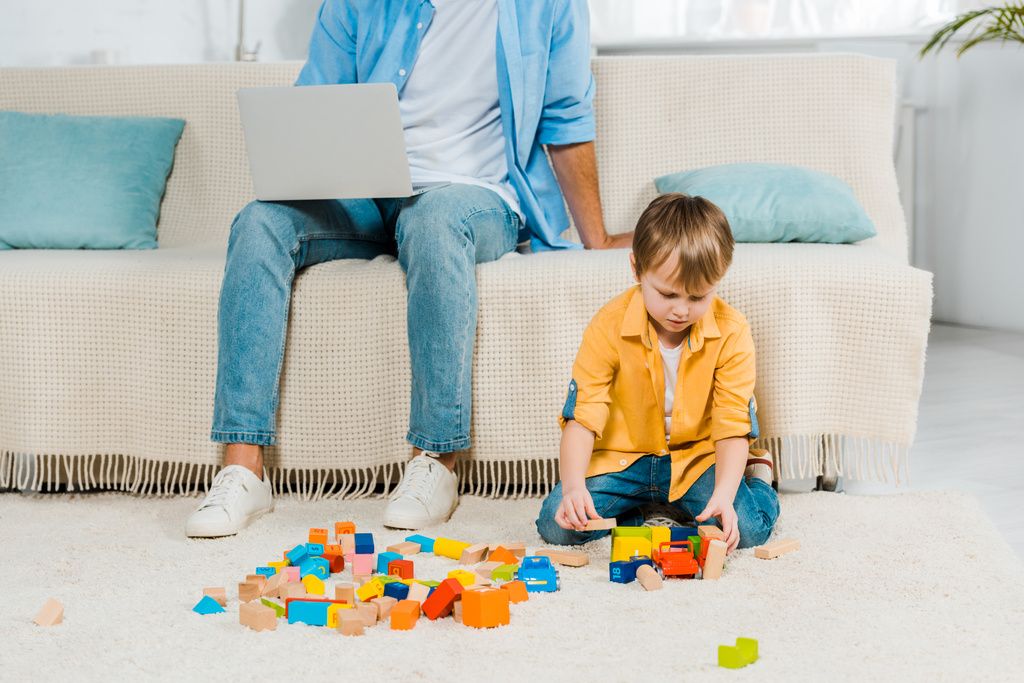 ο πατέρας κάθεται στον καναπέ και να χρησιμοποιούν φορητό υπολογιστή ενώ ο γιος preschooler παίζοντας με πολύχρωμα τουβλάκια στο σπίτι - Φωτογραφία, εικόνα