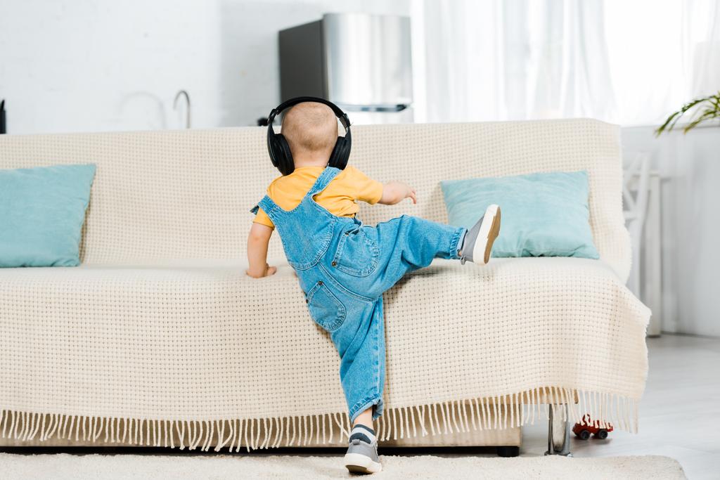 Rückansicht eines männlichen Kleinkindes mit Kopfhörer, das Musik hört und zu Hause auf der Couch klettert - Foto, Bild