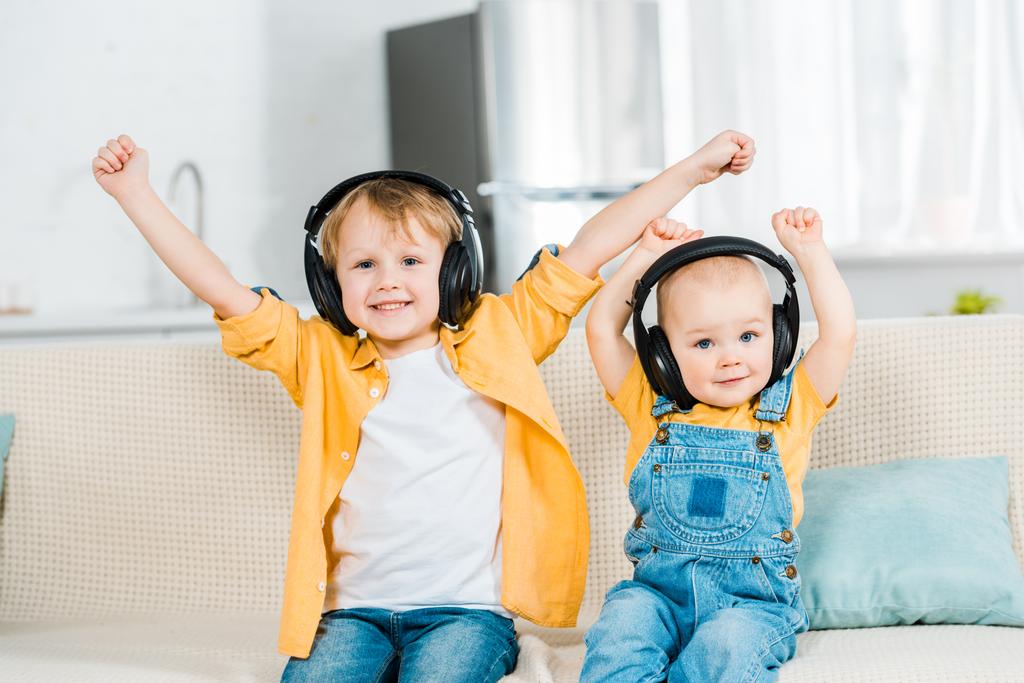 adorable brothers in headphones looking at camera and cheering with hands in air at home - Photo, Image