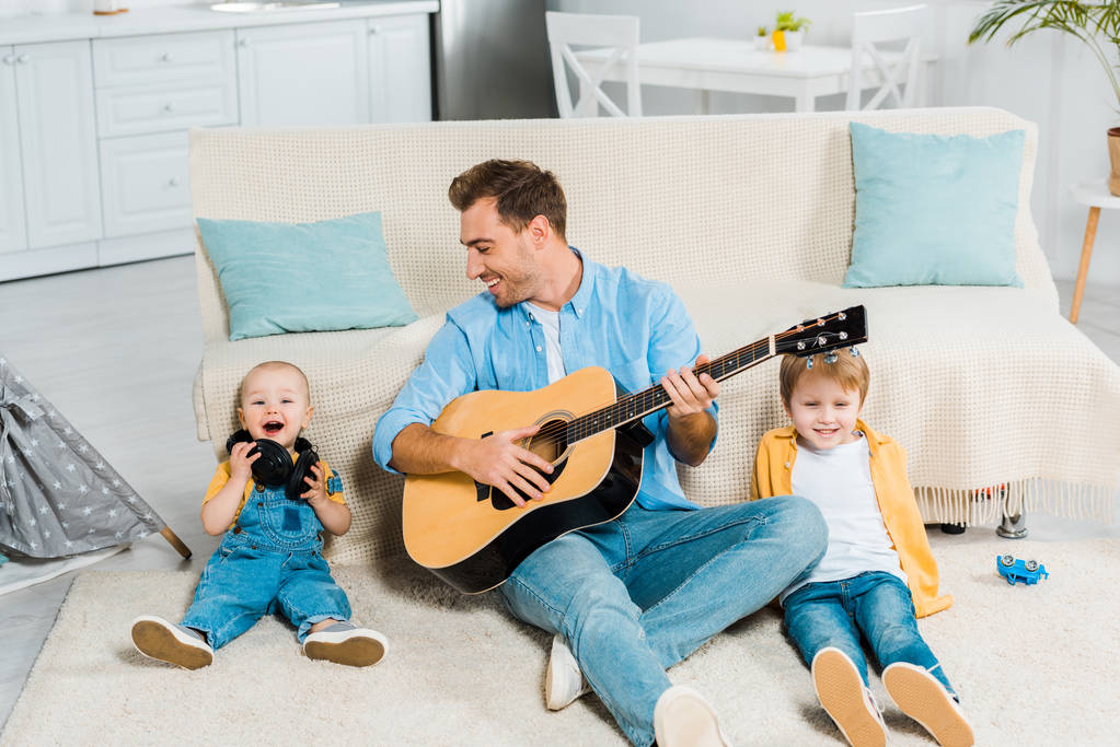 usmívající se otec hrál na akustickou kytaru rozkošný šťastný preschooler a batole synům doma - Fotografie, Obrázek