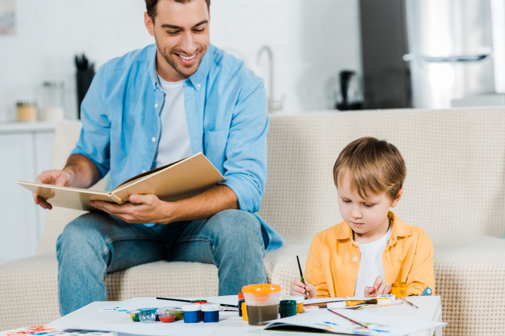 Lächelnder Vater liest Buch, während süßer Vorschulsohn zu Hause zeichnet - Foto, Bild