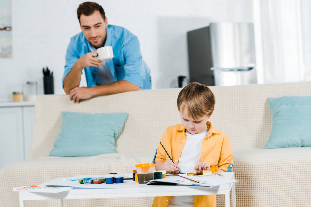 przedszkolak syn siedzi na kanapie i rysunek w domu, podczas gdy ojciec pije kawę na tle - Zdjęcie, obraz