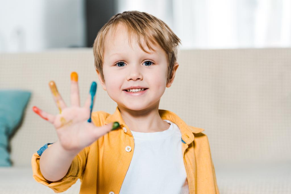 adorable smiling preschooler boy with colorful paint on hand looking at camera at home - Photo, Image