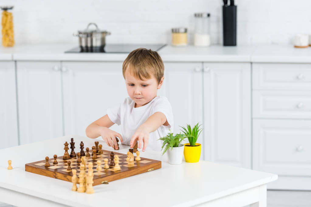очаровательный дошкольник сидит за столом и играет в шахматы дома
 - Фото, изображение