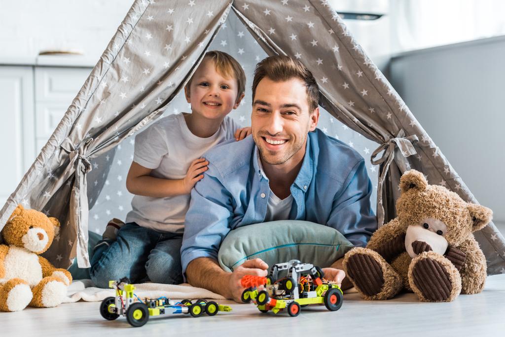 feliz padre e hijo preescolar con coches de juguete y osos de peluche mirando a la cámara mientras está sentado debajo de wigwam en casa
 - Foto, Imagen