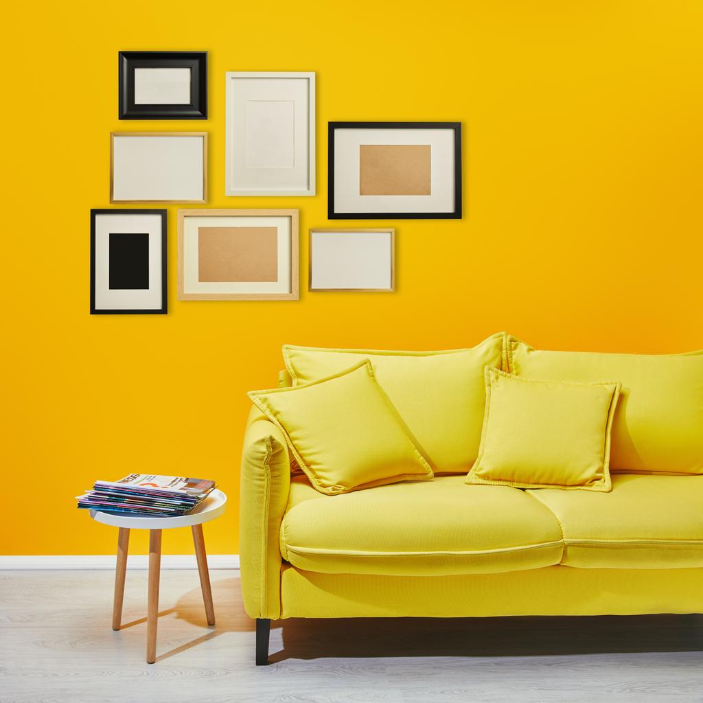 salontafel staande in de buurt van moderne gele Bank in de buurt van decoratieve frames opknoping op muur - Foto, afbeelding