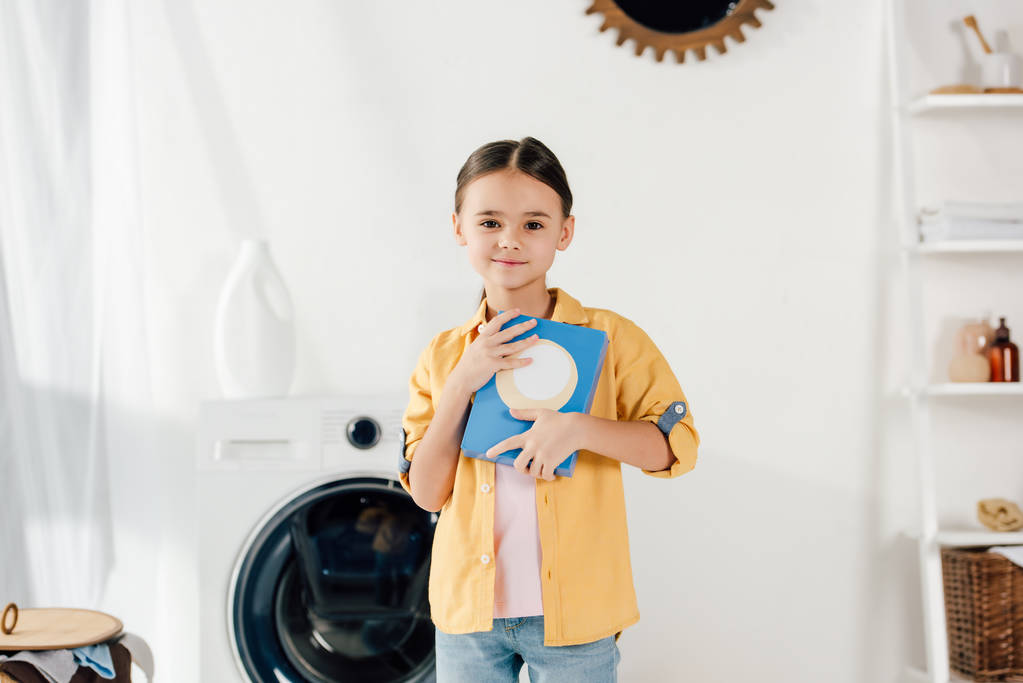 Kind steht neben Waschmaschine und hält Waschpulver in Waschküche - Foto, Bild