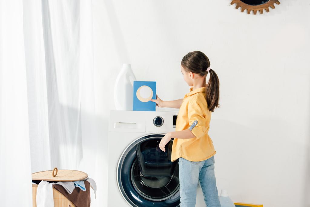 vue latérale de l'enfant en chemise jaune près de la laveuse prenant la poudre à laver dans la buanderie
 - Photo, image