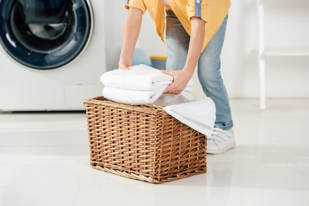 abgeschnittene Ansicht des Kindes in der Nähe der Waschmaschine, die Handtücher in den Korb in der Waschküche legt - Foto, Bild