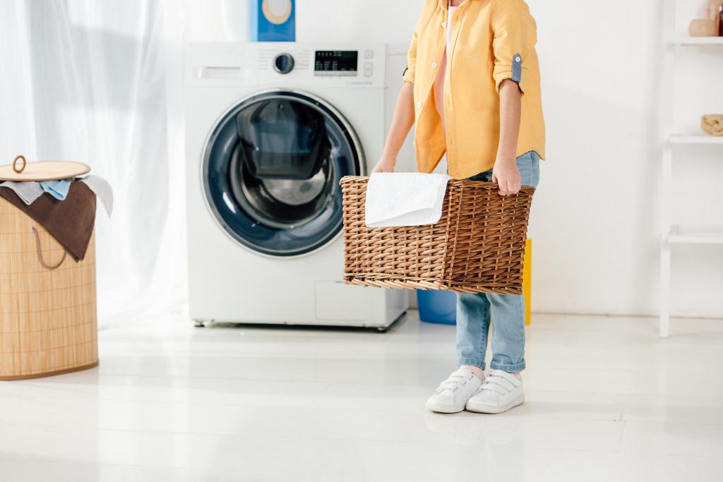 vista cortada da criança perto de lavadora segurando cesta na lavanderia
 - Foto, Imagem