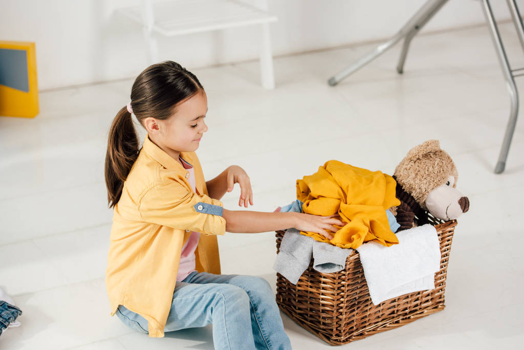 ребенок сидит и положить одежду в корзину на дому
 - Фото, изображение