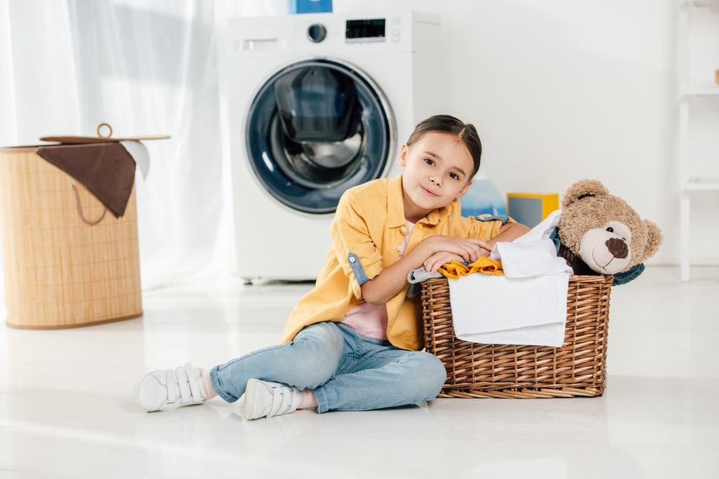 dziecko w żółte koszulki i jeansy siedzi w pobliżu kosz z niedźwiedź zabawka w pralni - Zdjęcie, obraz