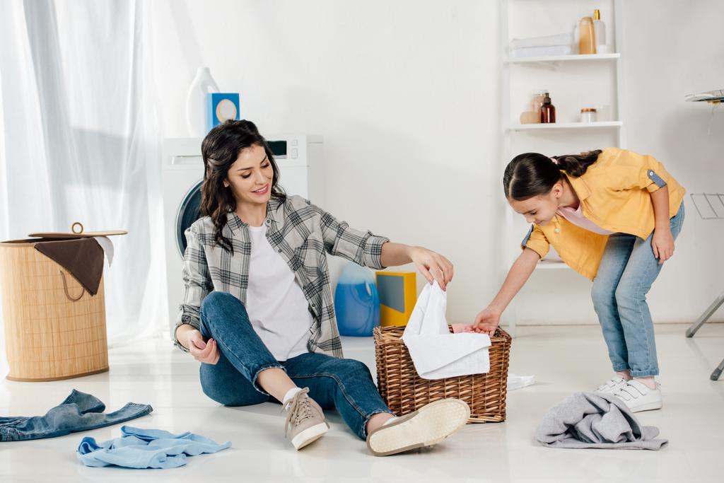 κόρη στο κίτρινο πουκάμισο και μητέρα βάζει ρούχα και πετσέτες στο καλάθι στο δωμάτιο πλυντηρίων - Φωτογραφία, εικόνα