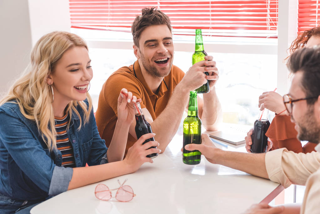 amis souriants tenant des bouteilles en verre avec de la soude et de la bière et parlant au café
  - Photo, image