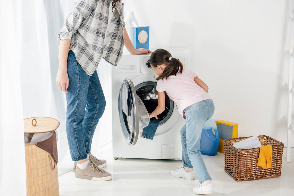 Tochter in rosa T-Shirt steckt Kleidung in Waschmaschine und Mutter steht in Waschküche - Foto, Bild