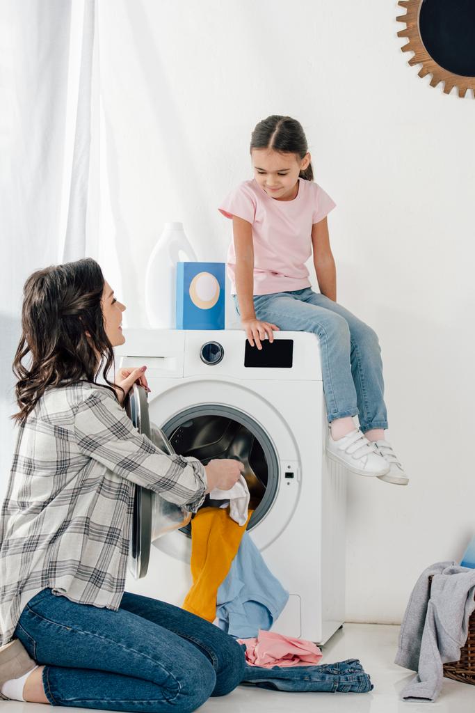 figlia in t-shirt rosa seduta sulla lavatrice madre astuta in camicia grigia che mette i vestiti in lavanderia
 - Foto, immagini
