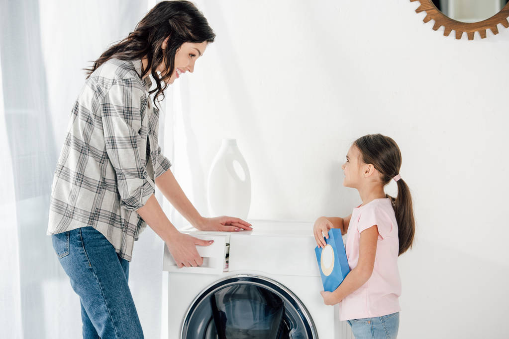 дочка в рожевій футболці тримає пральний порошок матері в сірій сорочці, відкриваючи пральну машину в пральній кімнаті
 - Фото, зображення