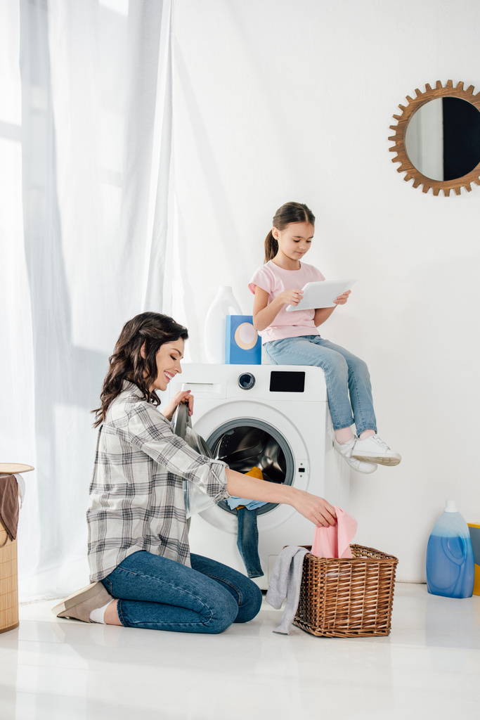 hija en camiseta sentada en la lavadora con tableta digital madre astuta en camisa gris tomando ropa de la cesta en la sala de lavandería
 - Foto, Imagen