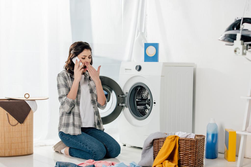Frau sitzt neben Waschmaschine, telefoniert mit Smartphone und gähnt in Waschküche - Foto, Bild