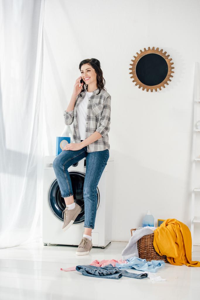 woman in grey shirt and jeans sitting on washer and talking on smartphone in laundry room - Photo, Image