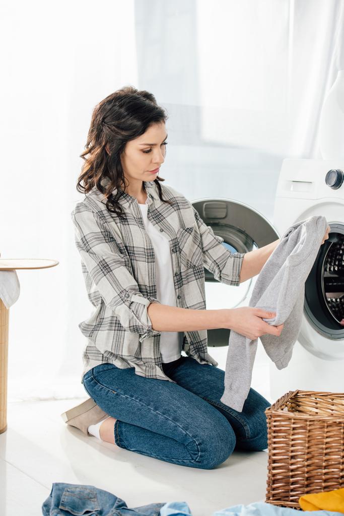 mujer en camisa gris y jeans sosteniendo la ropa cerca de la lavadora en la sala de lavandería
 - Foto, imagen
