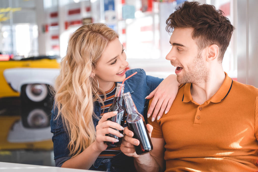 hermosa pareja sonriendo, abrazando y animando con botellas de vidrio en la cafetería
  - Foto, Imagen