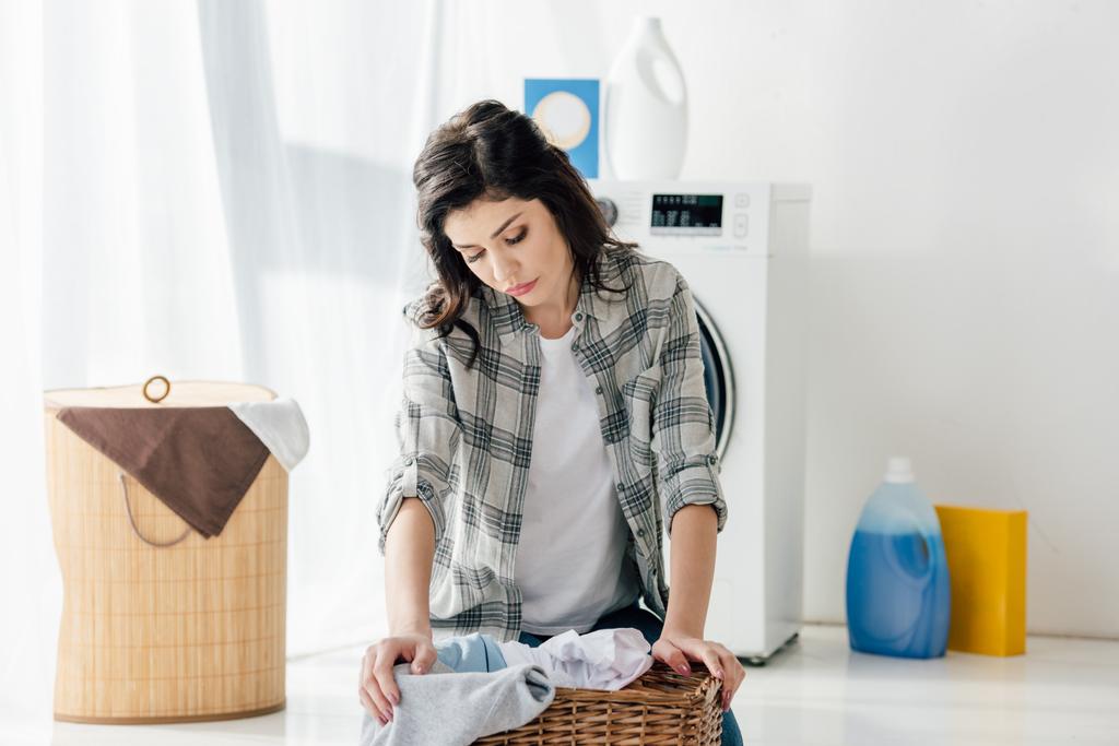 selective focus of upset woman in grey shirt sitting near basket in laundry room - Photo, Image
