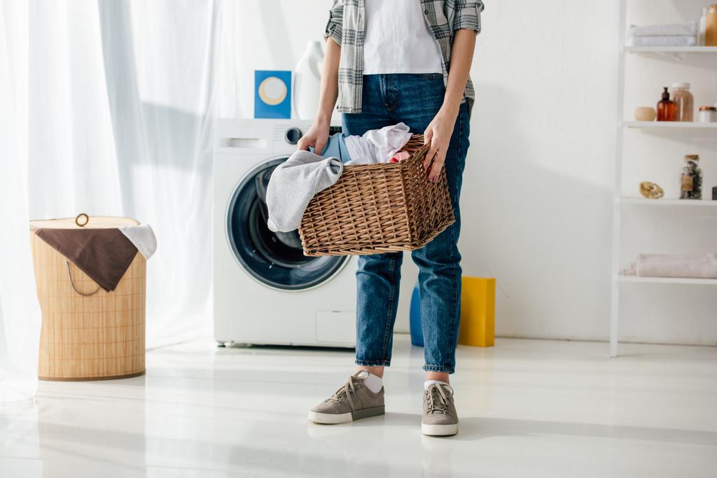 vista recortada de la mujer en camisa gris y jeans que sostienen la cesta en la sala de lavandería
 - Foto, imagen