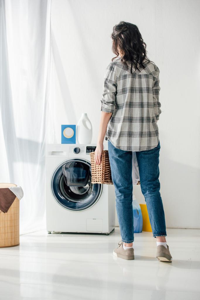 Rückansicht einer Frau in grauem Hemd und Jeans, die einen Korb neben der Waschmaschine in der Waschküche hält - Foto, Bild