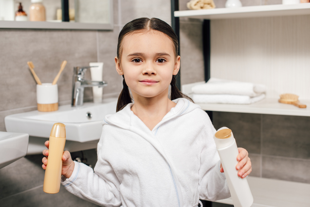 cute child in white bathrobe holding bottles with shower gel in bathroom  - Photo, Image