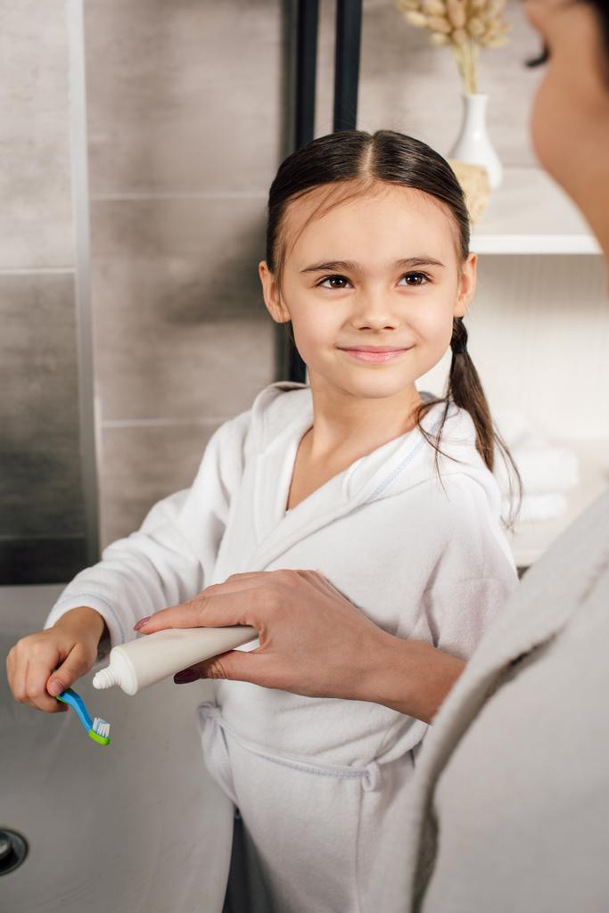 мать наносит зубную пасту из тюбика на дочь голубой зубной щетки в ванной комнате
 - Фото, изображение