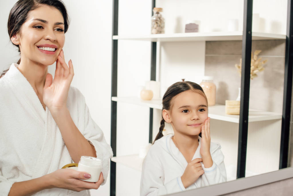 мать и дочь в белых халатах наносят косметический крем в ванной комнате
 - Фото, изображение