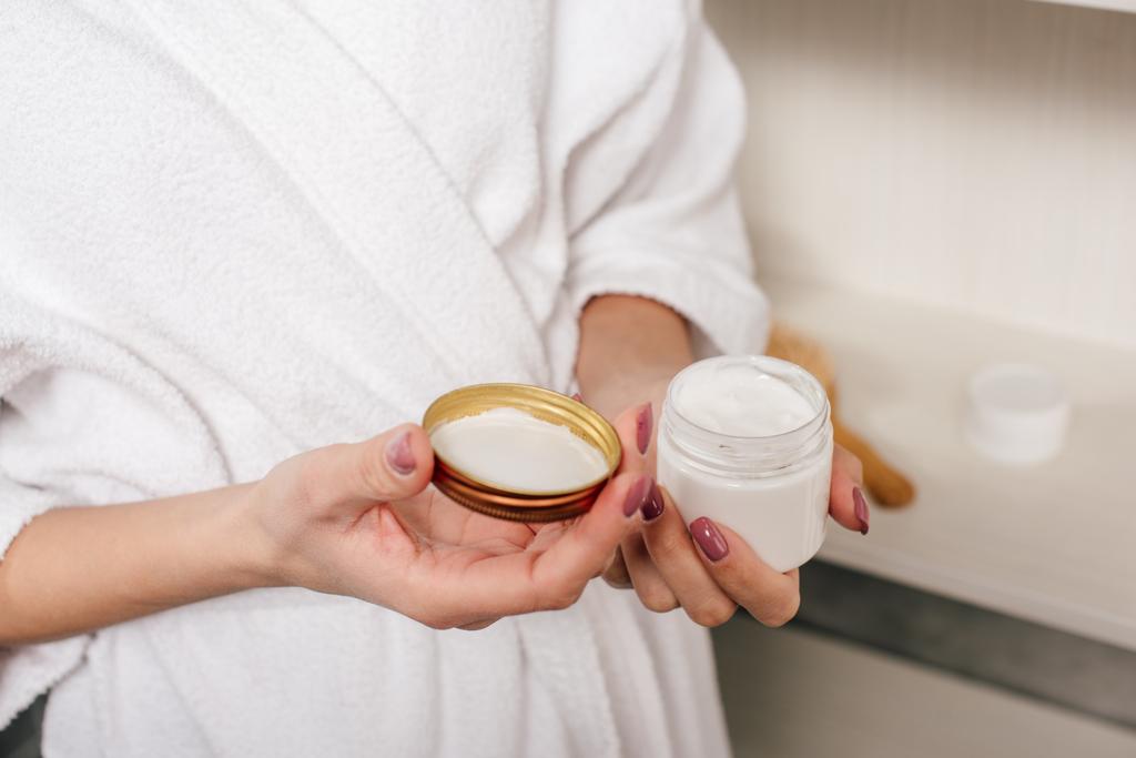 vista recortada de la mujer sosteniendo contenedor con crema cosmética en el baño
 - Foto, imagen