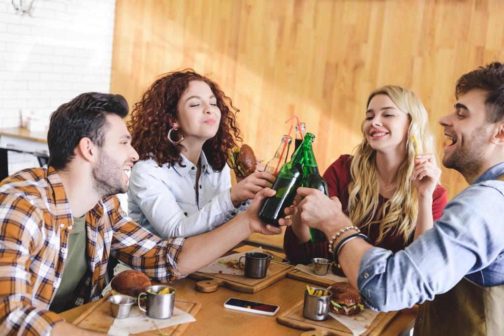 handsome and attractive friends smiling and cheering with glass bottles at cafe  - Photo, Image