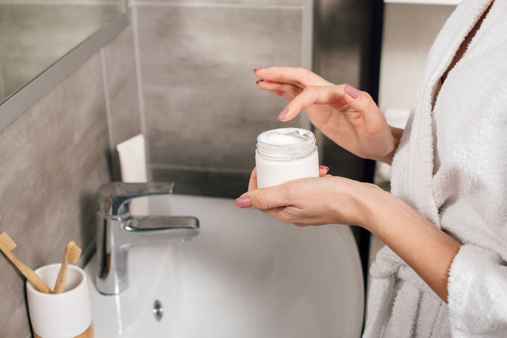 vista recortada de la mujer sosteniendo contenedor con crema cosmética en el baño
 - Foto, Imagen