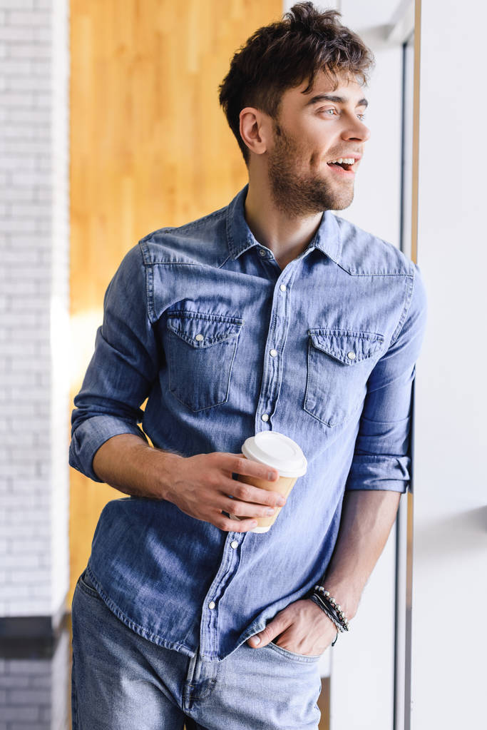 przystojny i uśmiechnięty człowiek stojący i trzymając kubek papierowy w kawiarni  - Zdjęcie, obraz