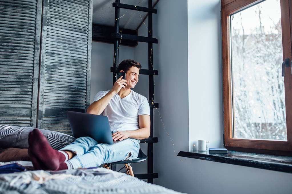 Dizüstü bilgisayarda çalışma ve evde smartphone üzerinde konuşurken genç adam - Fotoğraf, Görsel