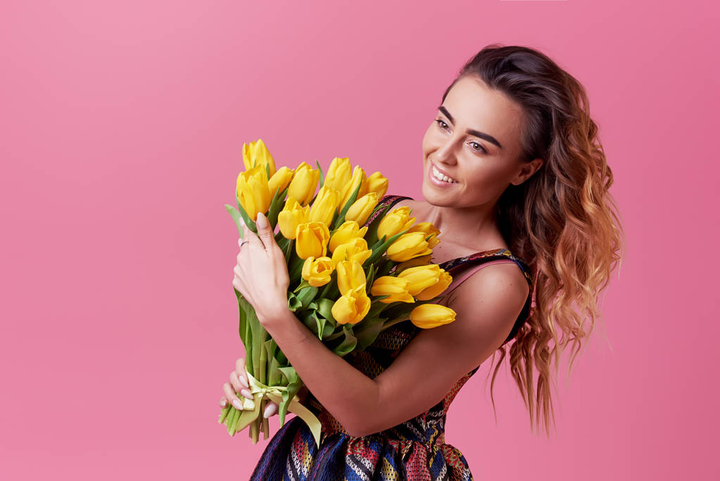 Vidám fiatal hölgy elegáns, divatos ruha, hogy izgatott, hogy a tavaszi sárga virágok-tulipán csokor a nőnap, elszigetelt, felett rózsaszín háttér.  - Fotó, kép