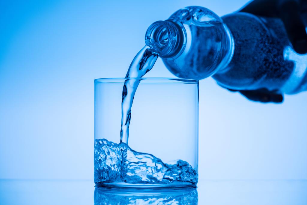 bijgesneden weergave van man stromende water uit plastic fles in glas op blauwe achtergrond - Foto, afbeelding