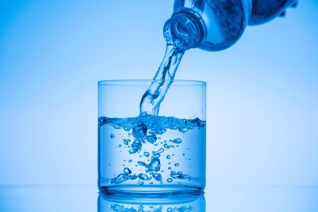 water pouring from plastic bottle in drinking glass on blue background - Photo, Image