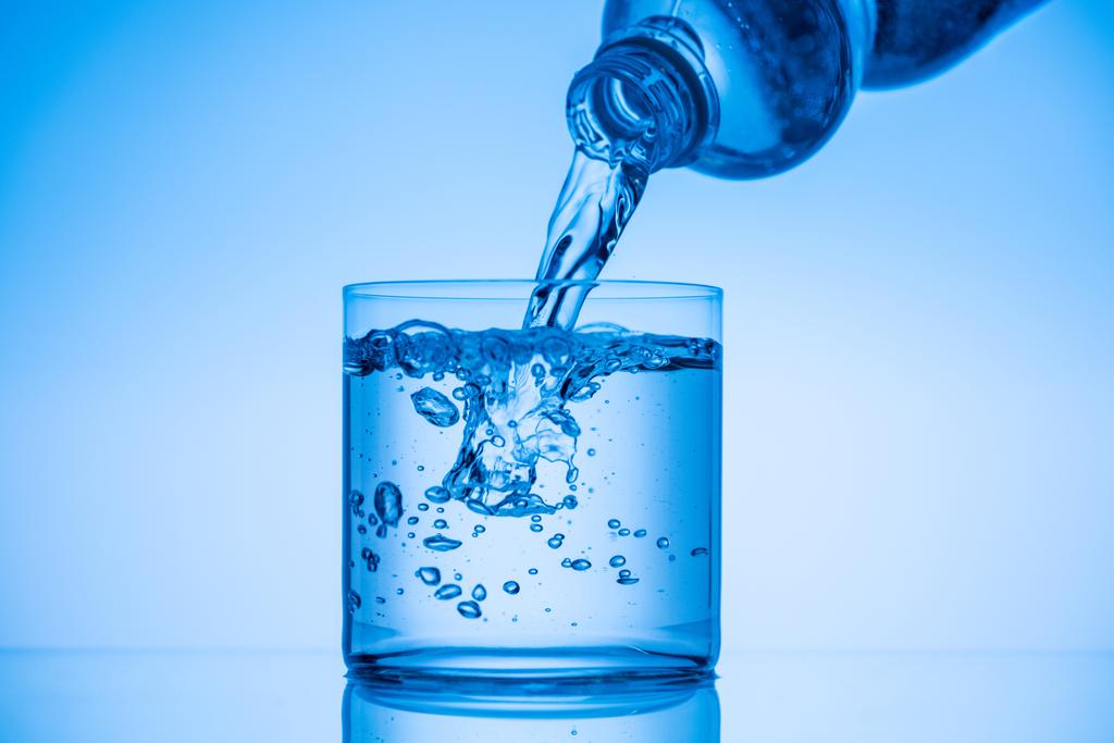 νερό ρίχνει από πλαστικό μπουκάλι στο γυαλί σε μπλε φόντο - Φωτογραφία, εικόνα