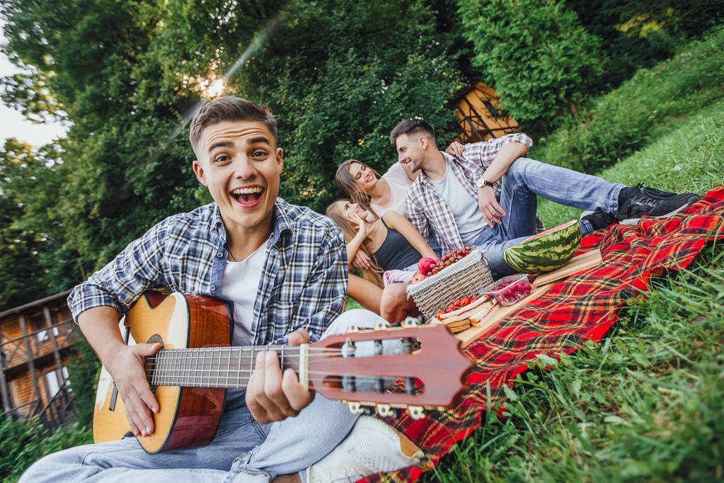 Uomo attraente seduto nell'erba e suonare la chitarra, picnic con tre amici
 - Foto, immagini