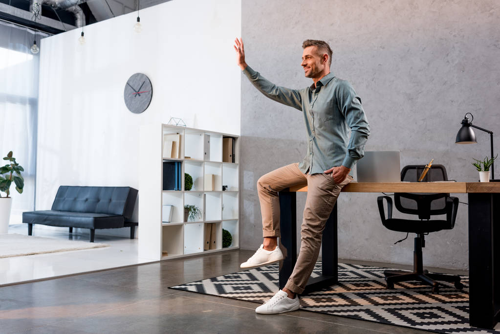empresário alegre acenando mão e de pé com a mão no bolso no escritório moderno
 - Foto, Imagem